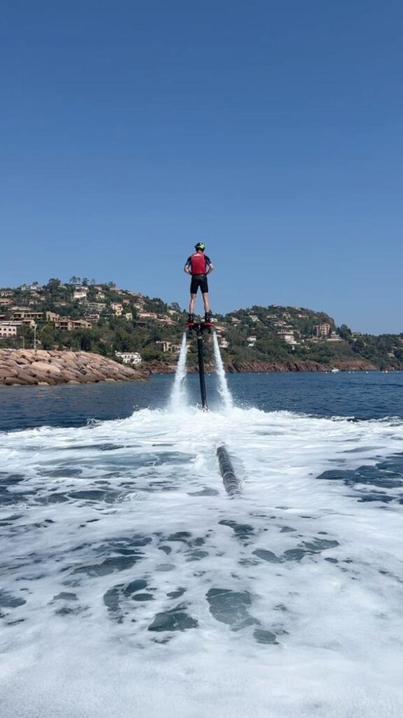 Flyboard au-dessus de la mer : l'aventure nautique ultime Flyboarder en gilet rouge survolant la mer, propulsé par des jets d'eau devant une côte résidentielle ensoleillée.