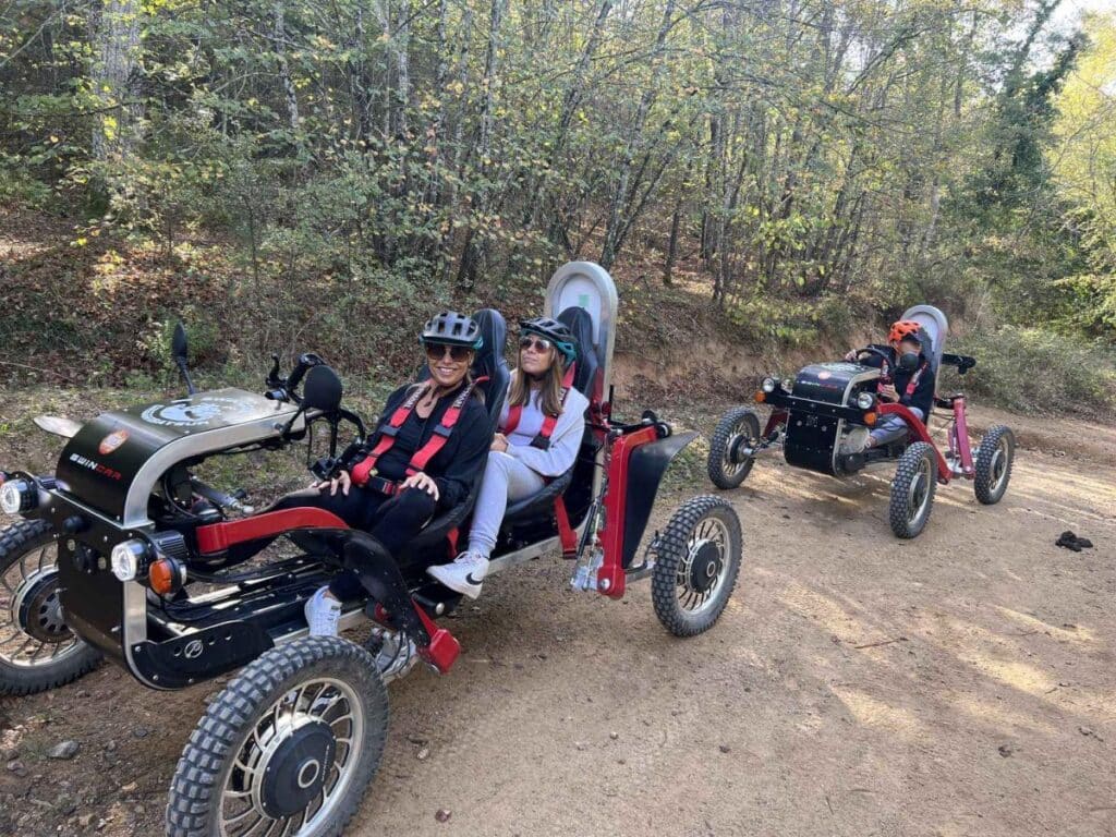 Deux femmes souriantes profitent d'une balade en Swincar électrique biplace sur un sentier forestier escarpé.