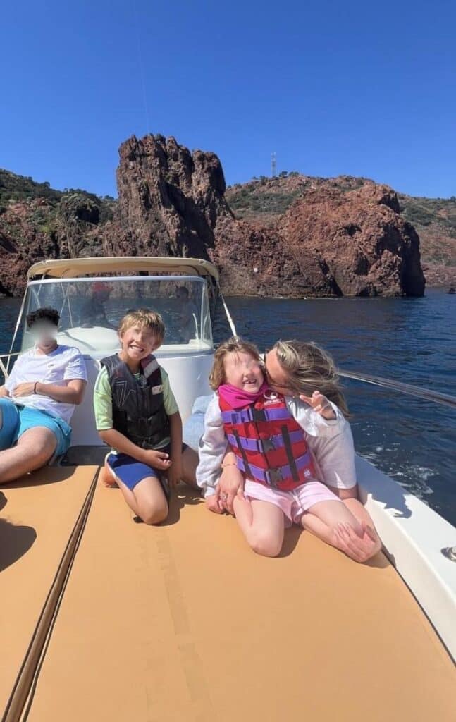 Balade en bateau en famille devant les falaises rouges Famille souriante sur un bateau devant des falaises rouges, mer d'un bleu profond et ciel dégagé.