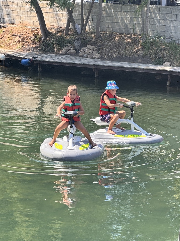 Enfants en Jet-Ski Gonflable : Activité Nautique Amusante Deux garçons en gilets rouges pilotent des mini jet-skis gonflables Geco et Jet Ski sur l'eau près d'un ponton en bois.