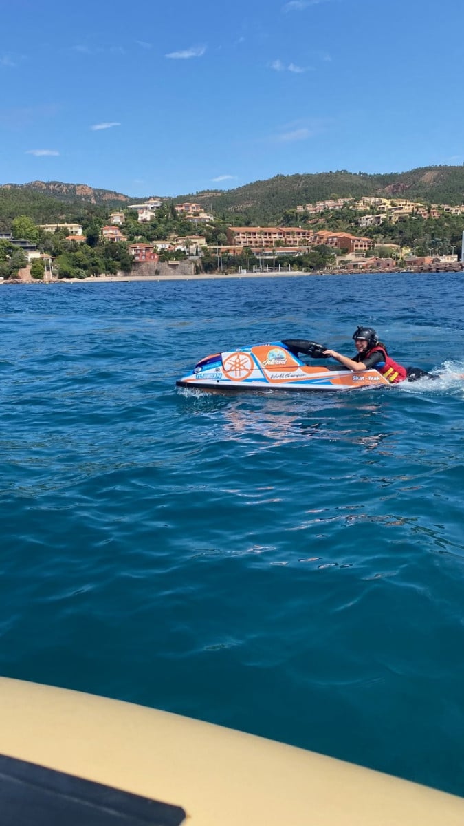 Personne pilotant un jet ski Yamaha orange et bleu sur une mer azur devant une côte vallonnée et ensoleillée.