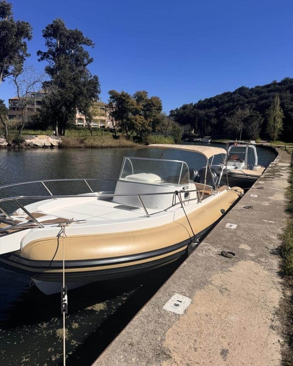 Bateau semi-rigide beige amarré au quai 19 le long d'un canal calme sous un ciel bleu dégagé.