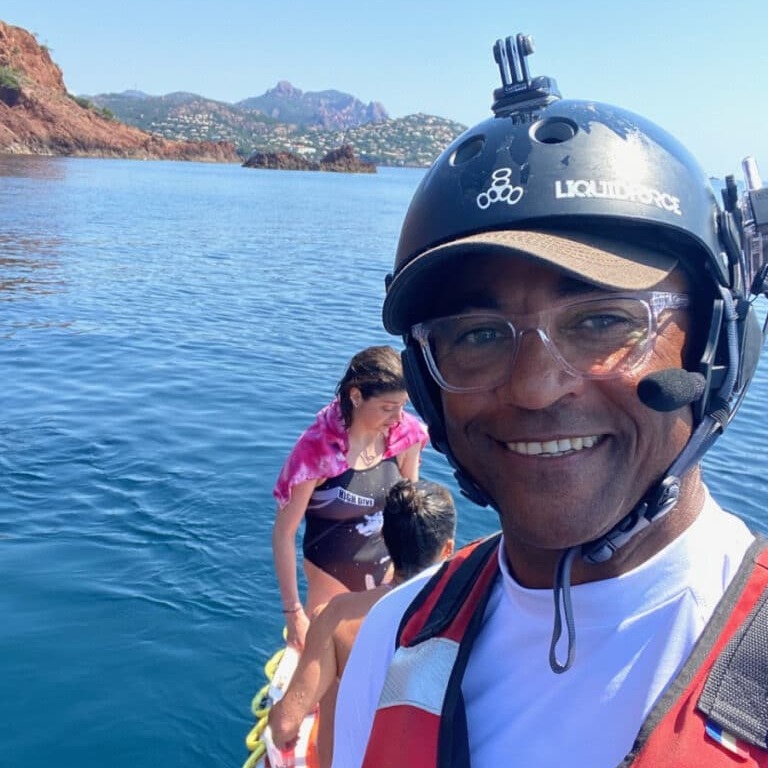 Activités nautiques en mer : Homme souriant avec casque Homme souriant avec casque Liquid Force et lunettes sur un bateau en mer devant une côte montagneuse.